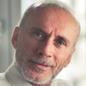 Man with gray hair and beard, wearing a light shirt and tie, seated near a window.